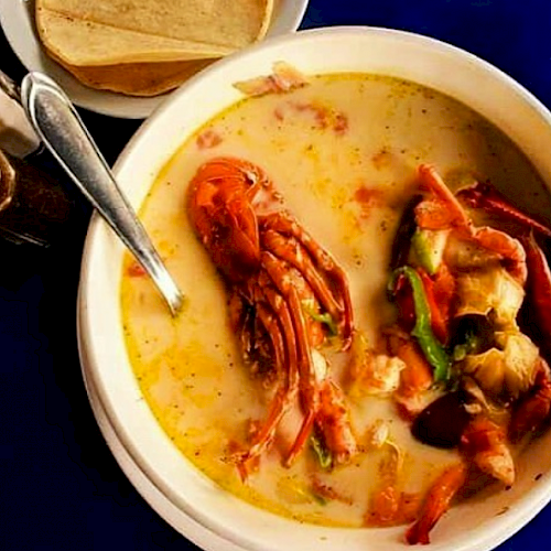 A bowl of seafood soup with various shellfish, a spoon, and tortillas on the side, placed on a blue surface.