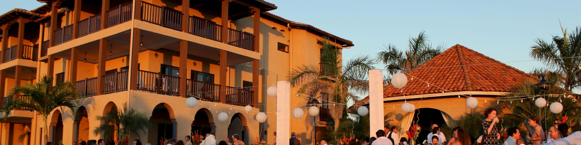 A group of people is seated at tables near a pool in front of a multi-story building during sunset, with festive decorations and palm trees.