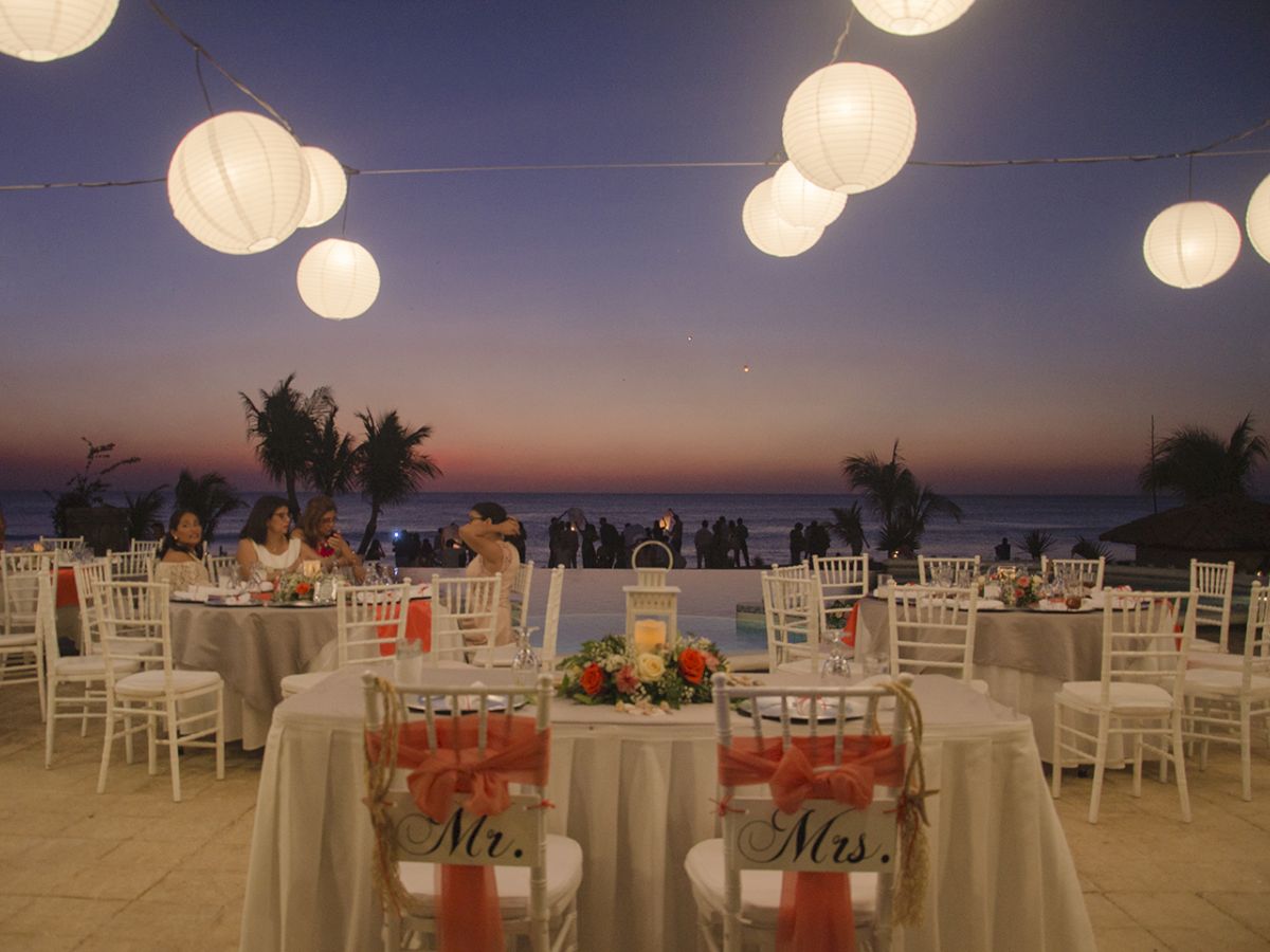 A beachside wedding reception at dusk, with lanterns hanging above, decorated tables, and chairs labeled "Mr." and "Mrs." in view.