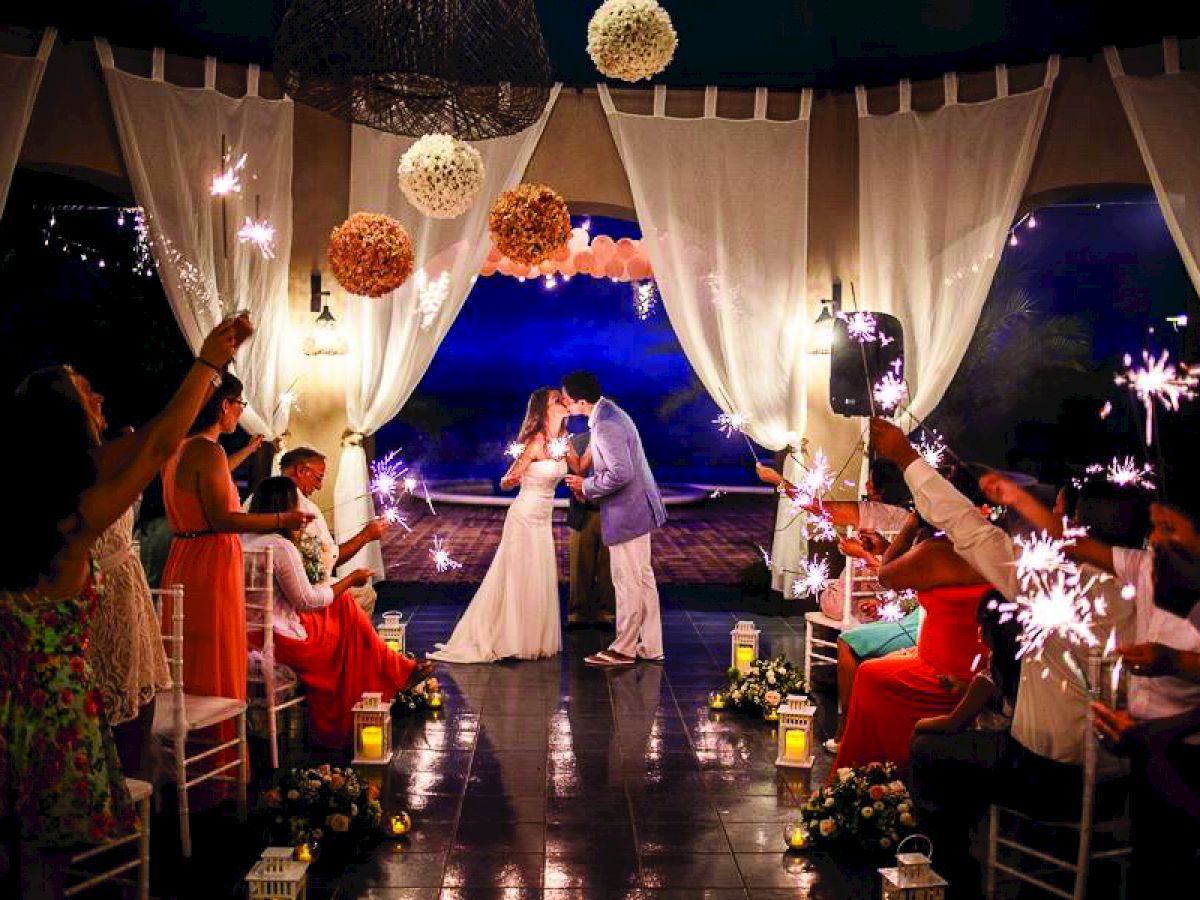 A newlywed couple shares a kiss at the alter under a decorated canopy, surrounded by guests holding sparklers in a romantic setting.