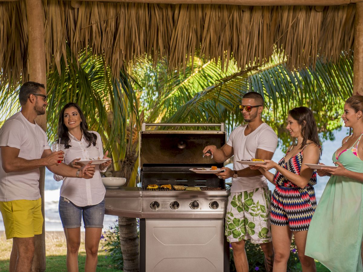 A group of five people enjoying a barbecue in a tropical outdoor setting with palm trees and a view of the ocean.