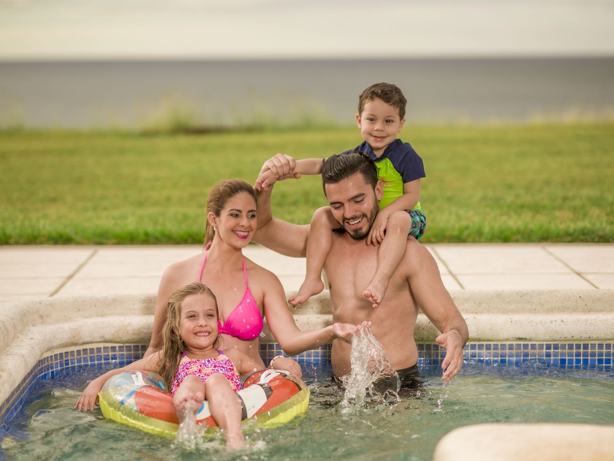 A family of four enjoys a pool with a grassy area and water in the background. The parents hold their children while smiling for the picture.