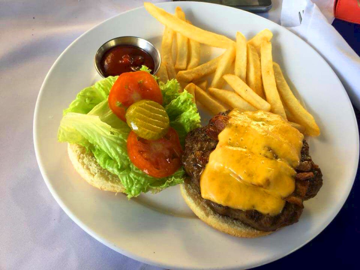 A cheeseburger with pickles, tomatoes, lettuce, and a side of fries on a white plate, accompanied by a small cup of ketchup.