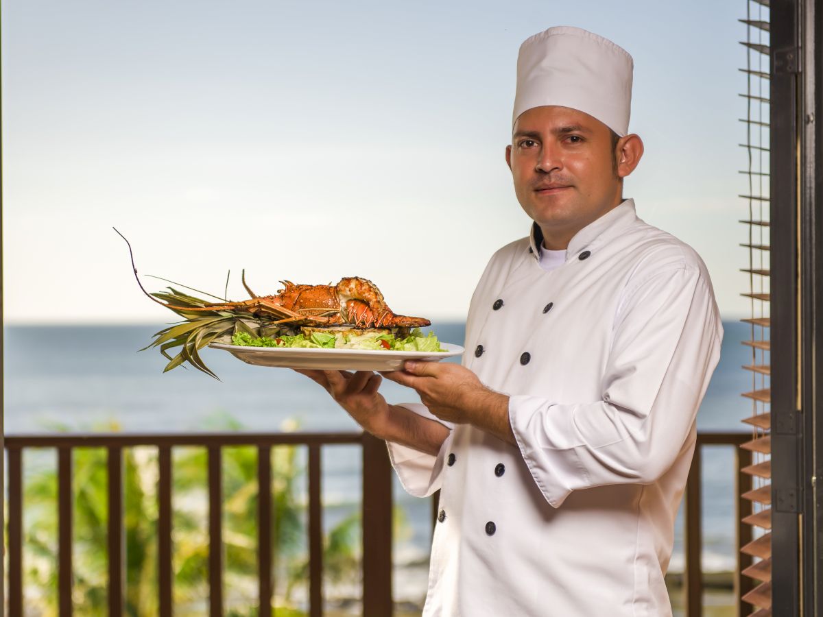 A chef dressed in a white uniform and hat holds a dish with seafood, standing in front of a balcony with a scenic ocean view in the background.