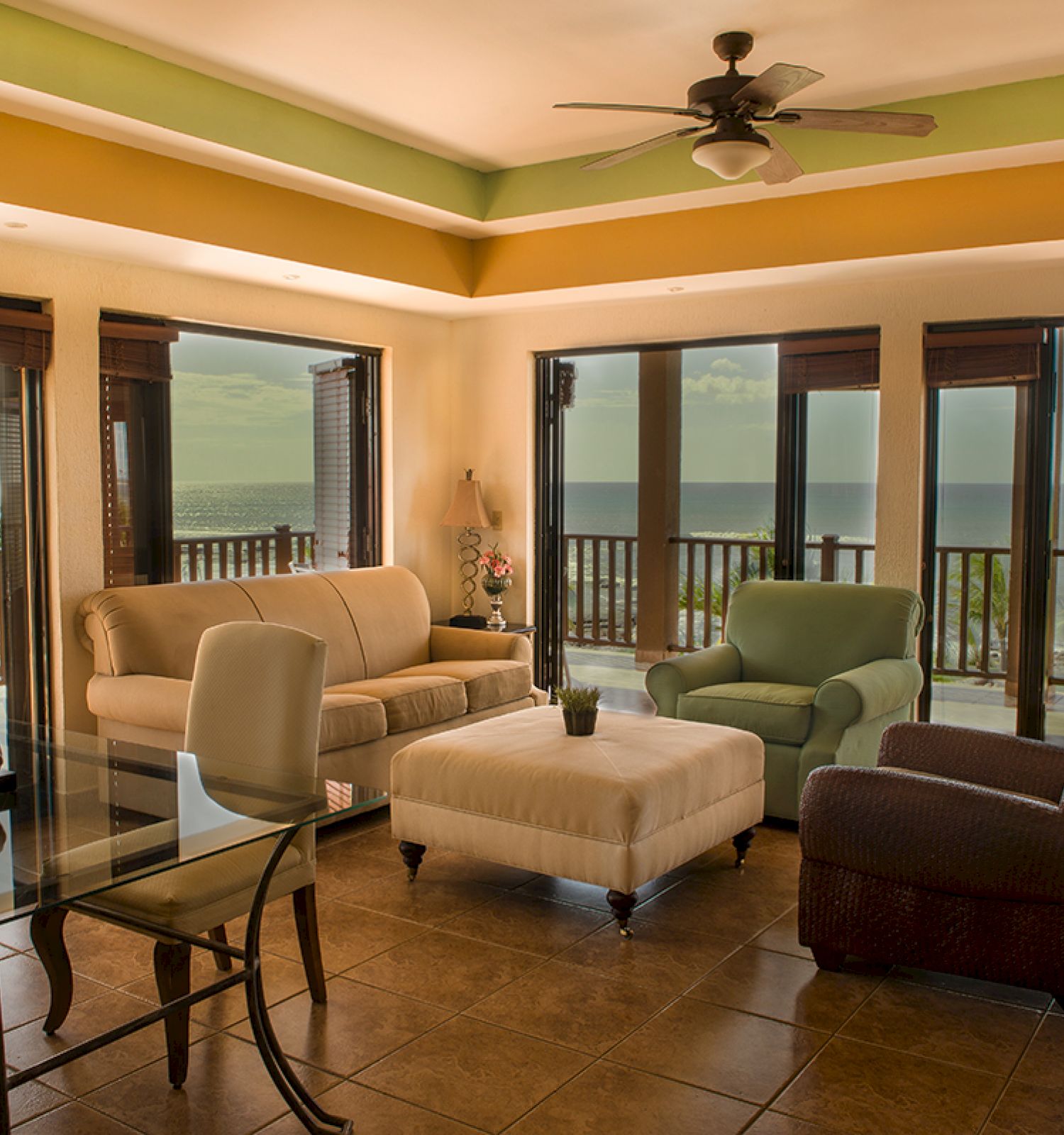 A spacious, elegant living room with multiple seating options, large windows showcasing an ocean view, and a ceiling fan completes the decor.