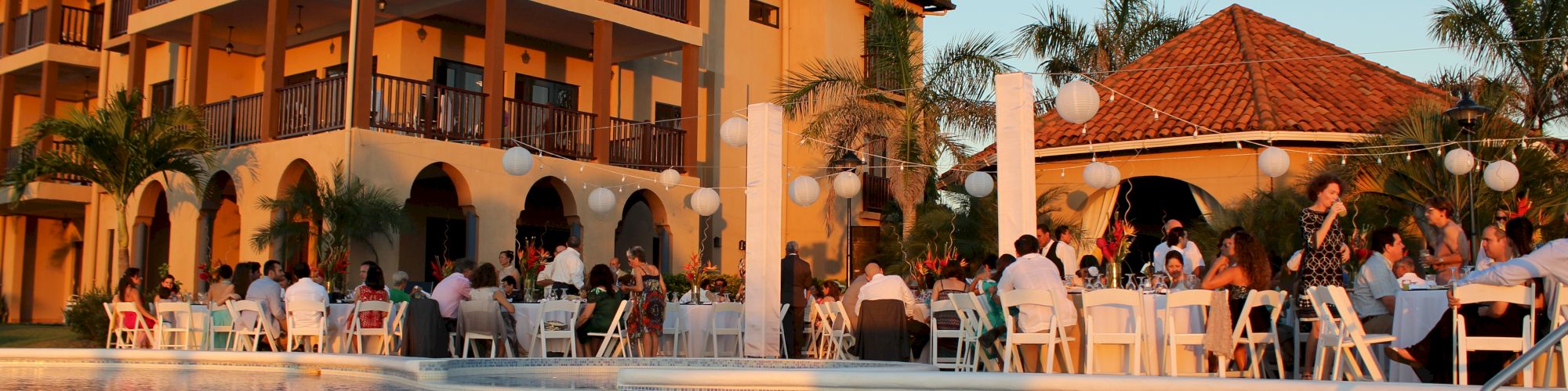 The image shows people gathered around tables next to a pool at a resort with Mediterranean-style buildings and palm trees in the background.