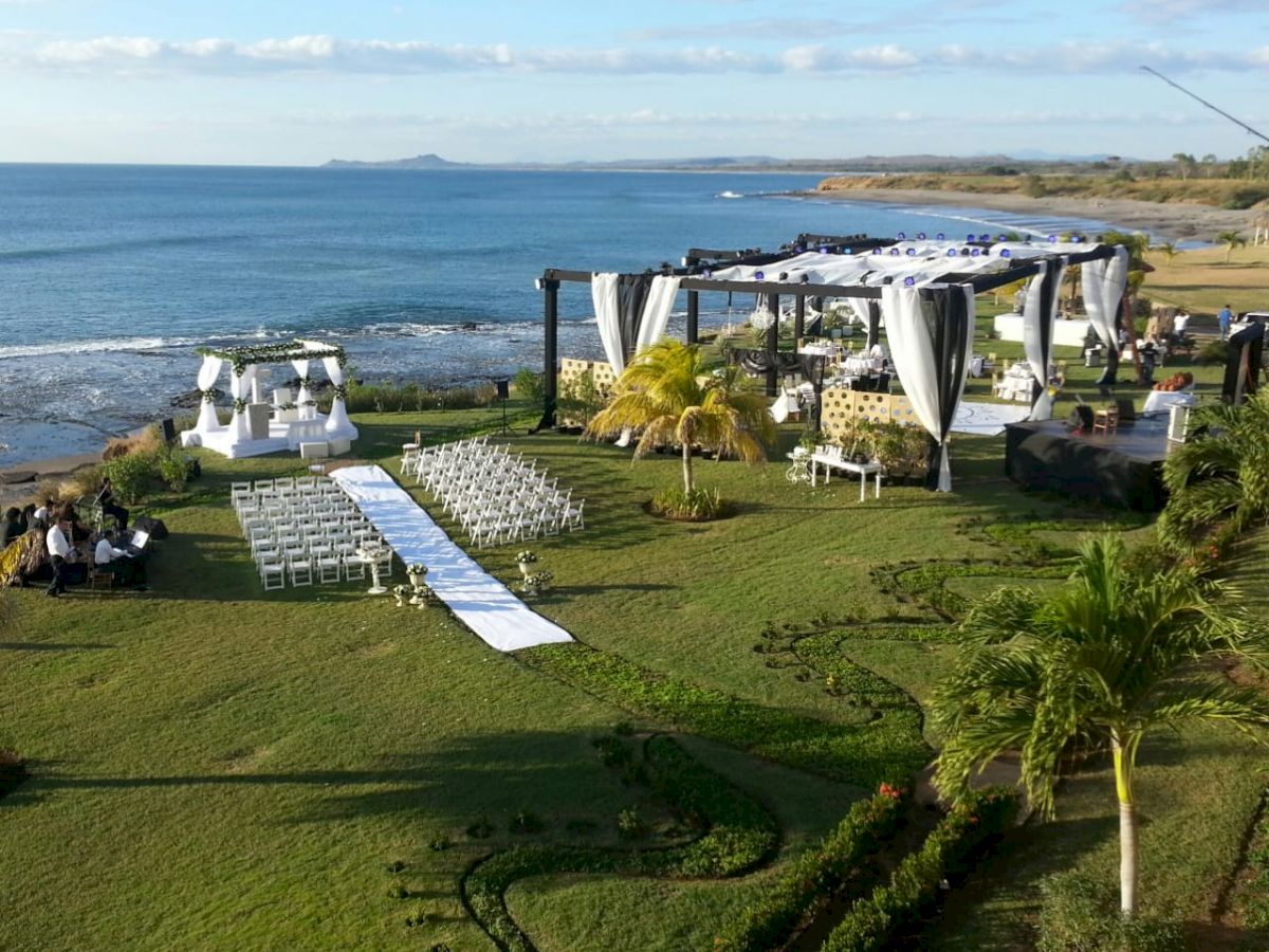 An outdoor wedding setup by the sea, featuring a white aisle, chairs arranged for guests, and decorated tents, with greenery and the ocean in the background.