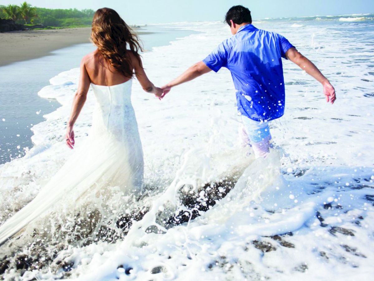 A couple is holding hands and running into the sea on a beach, with waves crashing around them. The woman is wearing a white dress.