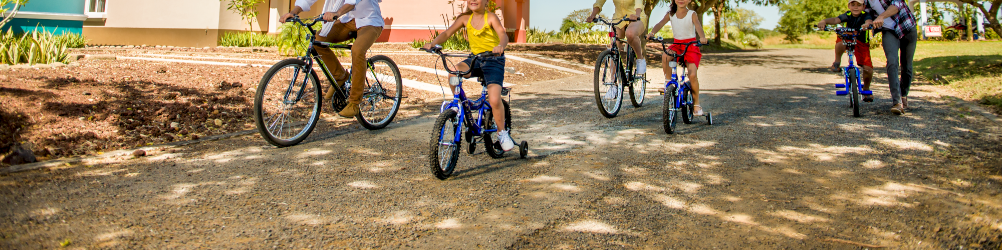 A group of people, including children, are riding bicycles on a sunny day along a suburban street lined with houses and trees, enjoying their time outdoors.