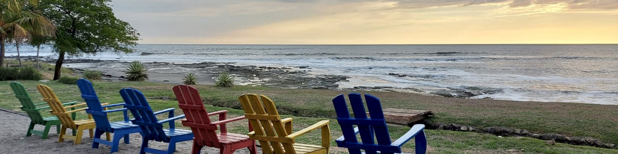 Several colorful Adirondack chairs face the ocean at a beach, with a sunset sky and waves on the shoreline in the background.