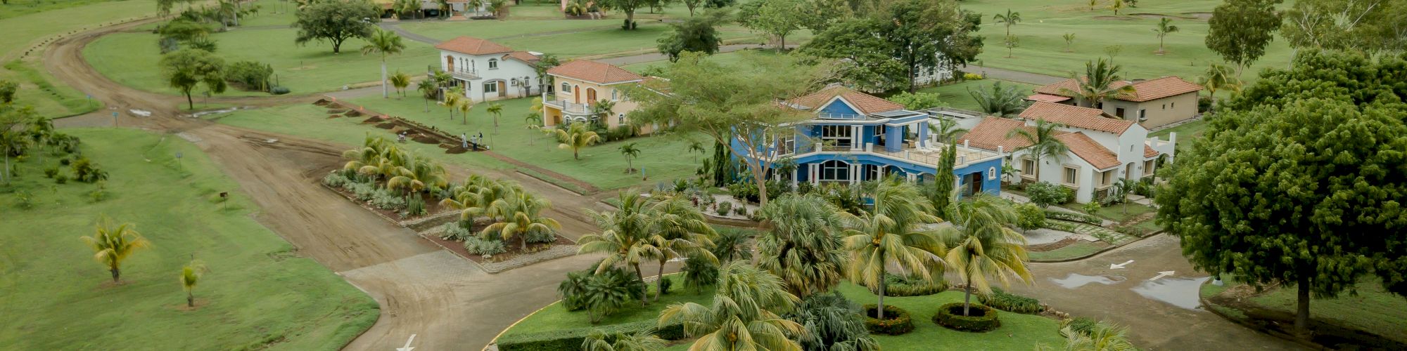 The image shows a scenic aerial view of a residential area with various houses, lush greenery, palm trees, and winding roads.