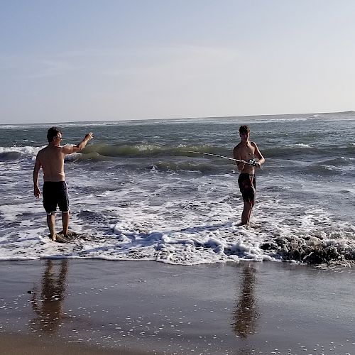 Two people are standing in the ocean waves on a sandy beach, one facing the water and the other gesturing. The sky is clear with calm waters.