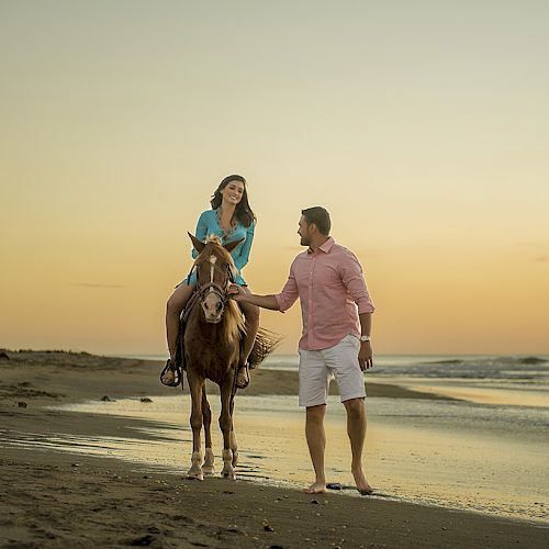 A woman riding a horse on the beach at sunset, with a man walking beside her and holding the reins, enjoying a peaceful moment together.