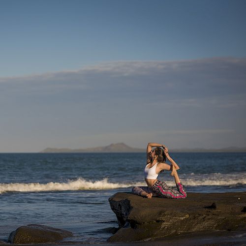 A person is practicing yoga on a rock near the ocean with waves in the background under a clear sky.