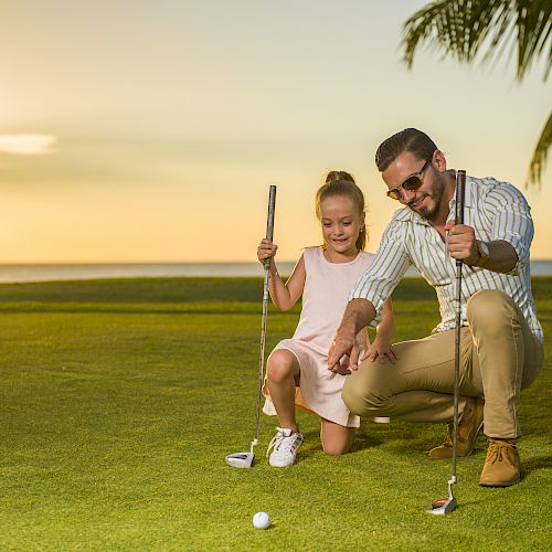 A man and young girl are on a golf course at sunset, holding golf clubs, with the man guiding the girl in playing a shot.