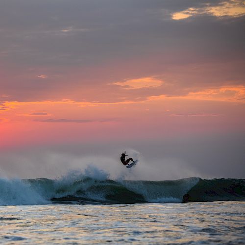 A surfer catches a wave at sunset, with vibrant colors in the sky and the sun low near the horizon. The waves crash amid the serene, picturesque scene.