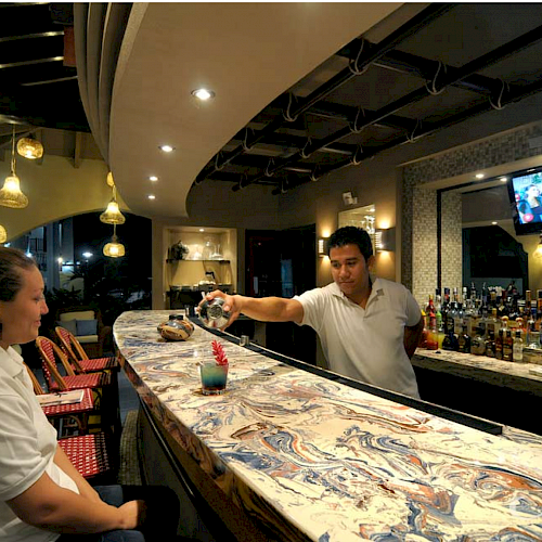 A bartender serves a drink to a seated customer at a bar, with a TV screen, bottles, and decorative lighting fixtures in the background.