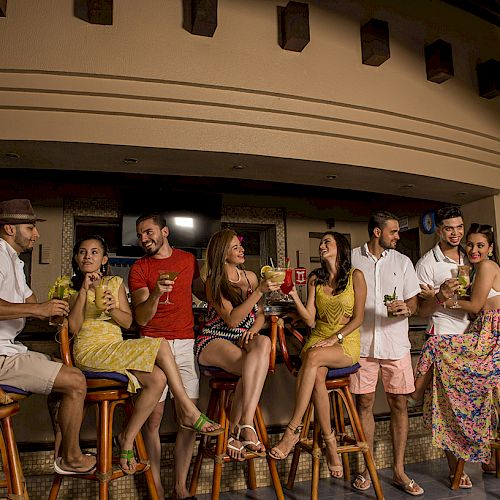 A group of people are socializing and holding drinks at an outdoor bar, all dressed in casual summer attire.