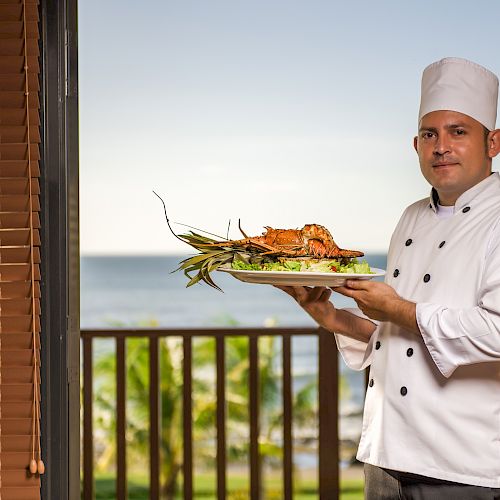 A chef in a white uniform and hat stands by a balcony with an ocean view, presenting a seafood dish. The background includes a railing and sea.