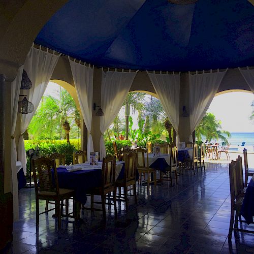 An open-air restaurant with a blue ceiling, surrounded by curtains, offers a view of the beach and lush greenery.