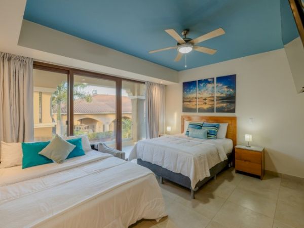 A modern bedroom featuring two beds with white linens, blue accent pillows, a ceiling fan, artwork on the wall, and a large window with a view.
