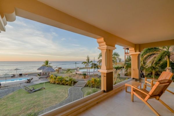 A beachfront patio with wooden lounge chairs overlooking a garden, pool, and palm trees facing the ocean at sunset.