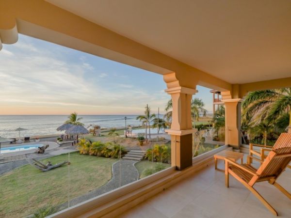 A beachfront patio with wooden lounge chairs overlooking a garden, pool, and palm trees facing the ocean at sunset.