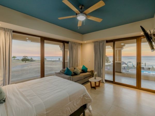 A modern bedroom with large windows offers a view of the sunset over the ocean, a bed, a sofa, a ceiling fan, and a mounted TV.