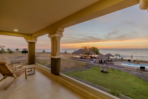 A beachfront balcony view showcasing lounge chairs, a pool area with thatched huts, and a serene sunset over the ocean.