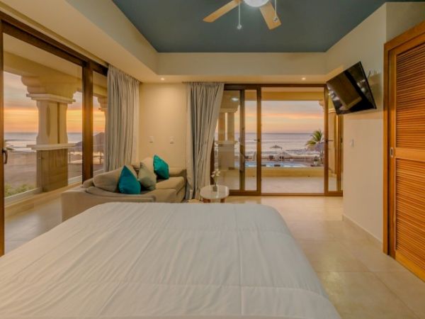 A cozy bedroom with a modern design featuring a bed, a small seating area, a wall-mounted TV, and large windows offering an ocean view at sunset.