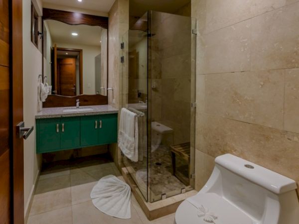The image shows a bathroom with a toilet, glass-enclosed shower, green vanity with sink, mirror, and beige tiled walls and floor.