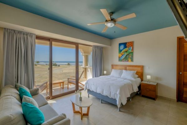 A cozy, modern bedroom with a large bed, sofa, ceiling fan, balcony, and ocean view. Artwork on the wall adds a pop of color.