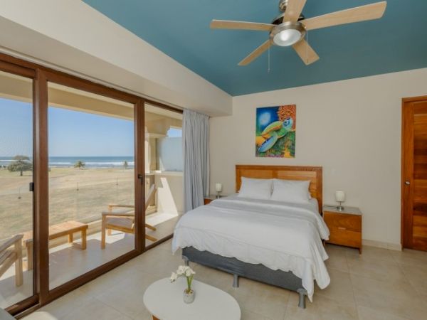 A bright bedroom with ocean view, features a double bed, wooden furniture, ceiling fan, and wall art, opening to a balcony with seating.