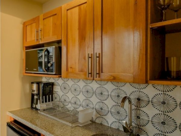 A kitchen with wooden cabinets, a microwave, coffee maker, dish rack, and a patterned tiled backsplash, ending the sentence.