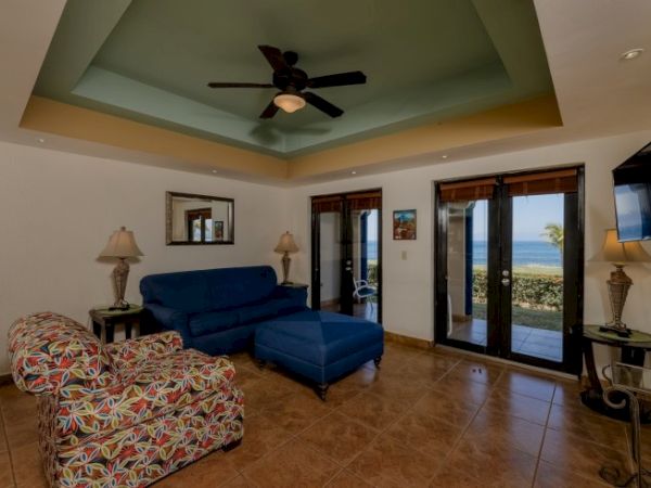 A cozy living room with colorful furniture, ceiling fan, and ocean view visible through glass doors, with tiled floors and wall decorations.