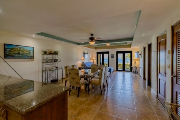 The image shows a modern, spacious living and dining area with a tiled floor, comfortable seating, artwork, and large glass doors leading outside.