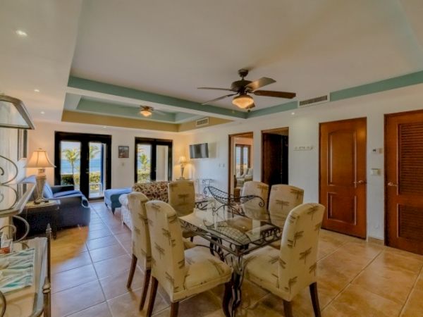 A well-furnished living area with a dining table, chairs, ceiling fans, and doors leading to a balcony or patio with a view of the outdoors.