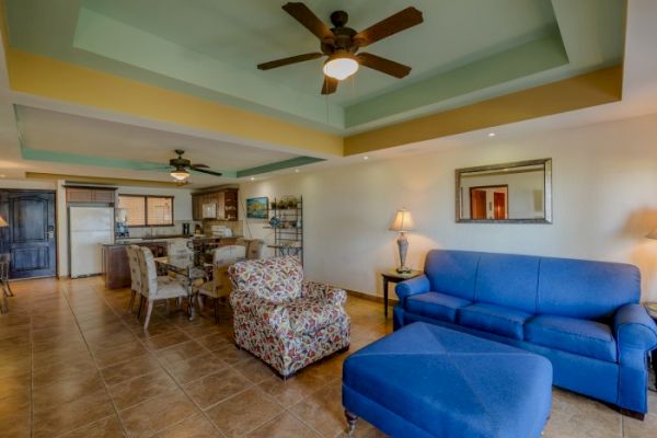 A modern living room with blue and patterned seating, a ceiling fan, open kitchen, and dining area, with warm lighting and tile flooring.