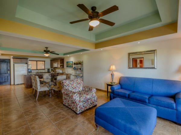 A modern living room with blue and patterned seating, a ceiling fan, open kitchen, and dining area, with warm lighting and tile flooring.