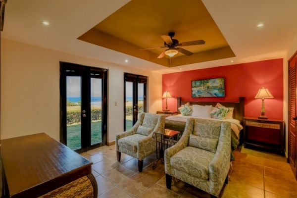 This image shows a cozy bedroom with a king-size bed, two armchairs, a ceiling fan, and a view of the outdoors through glass doors, warmly lit by lamps.