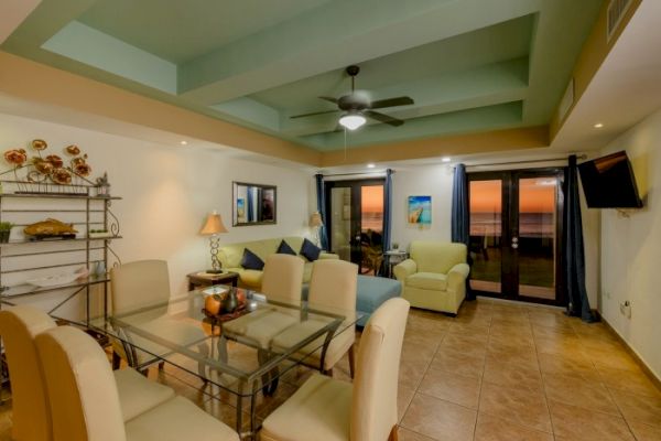 A cozy living and dining area with a glass table, beige chairs, sofa, armchair, ceiling fan, wall-mounted TV, and glass doors showing a sunset view.