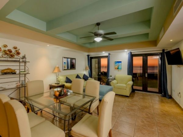 A cozy living and dining area with a glass table, beige chairs, sofa, armchair, ceiling fan, wall-mounted TV, and glass doors showing a sunset view.