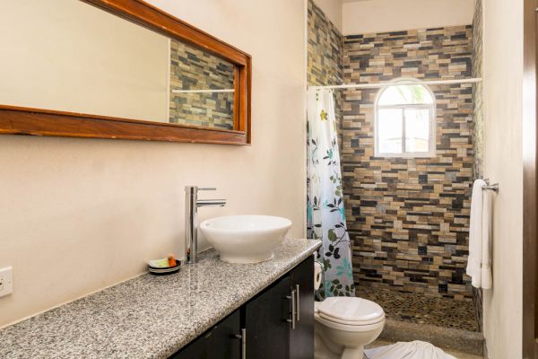 The image shows a modern bathroom with a granite countertop, vessel sink, large mirror, toilet, shower with a tiled wall, and light-colored floor tiles.