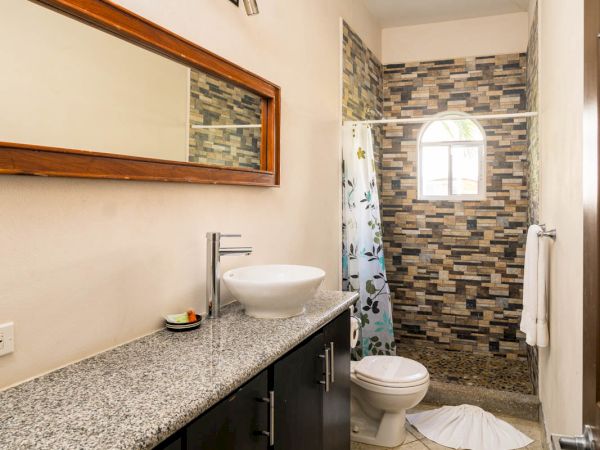The image shows a modern bathroom with a granite countertop, vessel sink, large mirror, toilet, shower with a tiled wall, and light-colored floor tiles.