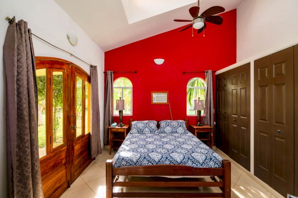 A cozy bedroom with a wooden bed, blue patterned bedding, red accented wall, twin bedside tables, large wooden doors, and a ceiling fan.