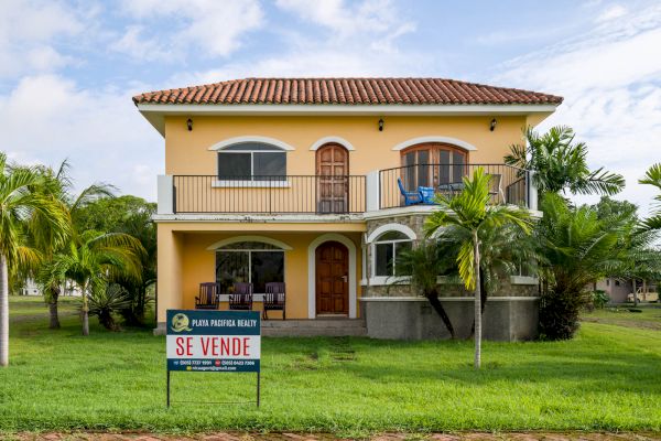 A two-story yellow house with balconies and a tiled roof is for sale. There are palm trees in the yard and a 