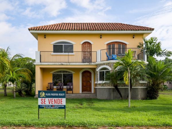 A two-story yellow house with balconies and a tiled roof is for sale. There are palm trees in the yard and a "Se Vende" sign in front.