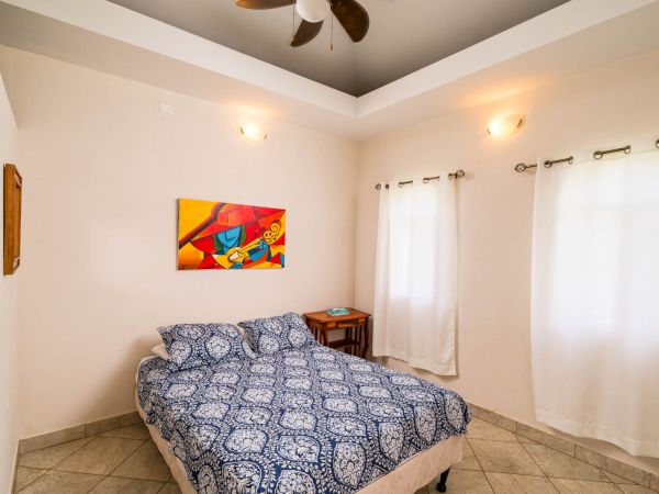 A cozy bedroom with patterned bedspread, two windows with curtains, colorful wall art, and a ceiling fan above.