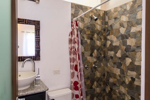 A bathroom with a toilet, sink basin, mirror, and a stone-tiled shower area with a red shower curtain featuring heart patterns ends the sentence.