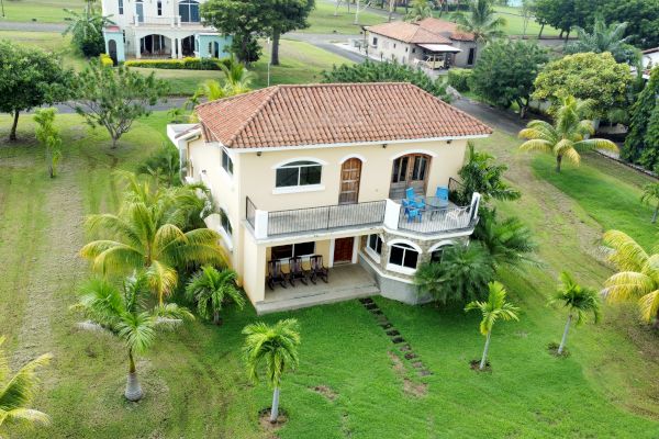 An aerial view of a Mediterranean-style house with a red-tiled roof, balcony, and lush greenery with palm trees surrounding it.